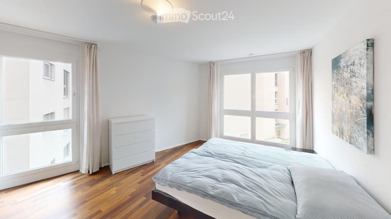 Bright bedroom with wooden flooring, large windows with curtains, a bed with grey bedding, a radiator, and a modern abstract artwork on the wall.