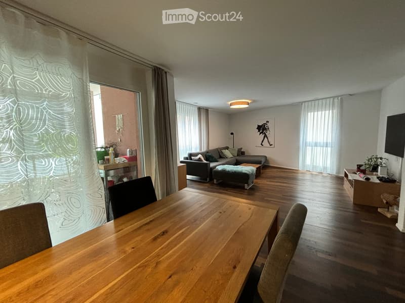 Open-plan living and dining area with wooden flooring, large windows, a grey sofa, coffee table, ottoman, dining table with chairs, and a TV screen.