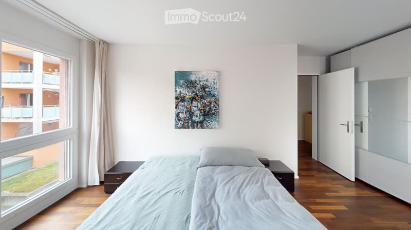 Modern bedroom with a single bed, nightstands, a colorful abstract painting on the wall, and a large window offering a view of the balcony and surrounding buildings.
