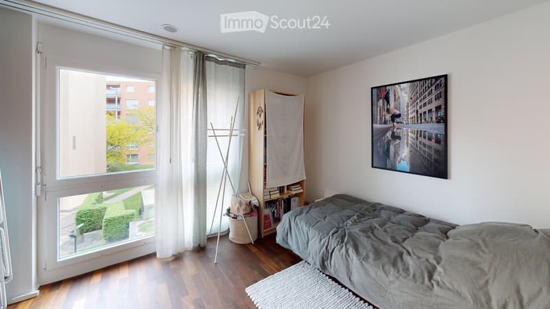 Modern bedroom with wooden flooring, large window with view, bed with grey duvet, bookshelf, picture frame, and coat rack.