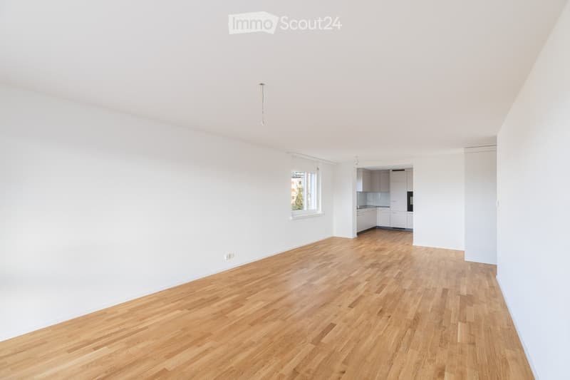 Empty room with light-colored wooden flooring and white walls, featuring a window and an adjacent kitchen area with white cabinets and appliances.