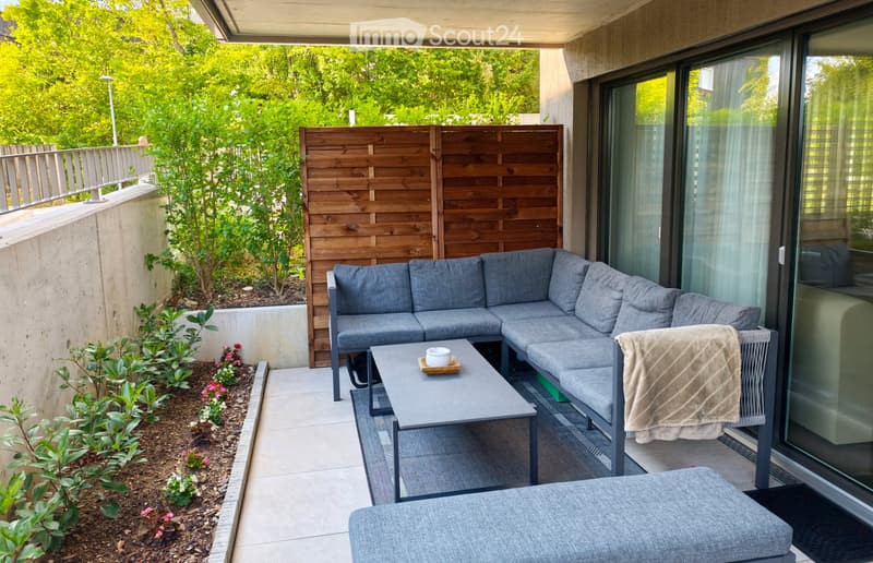 Modern balcony with grey sectional sofa, coffee table, and a small chair with a blanket draped over it. Wooden lattice screen and small garden area with plants and flowers.