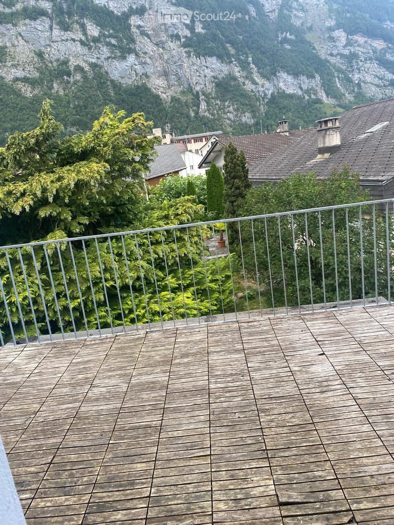 Wooden deck with metal railing, overlooking a lush green area with trees and distant mountains.