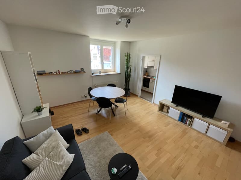 Modern living room with wooden flooring, a sofa, coffee table, dining table with chairs, TV, and a small kitchen area visible in the background.