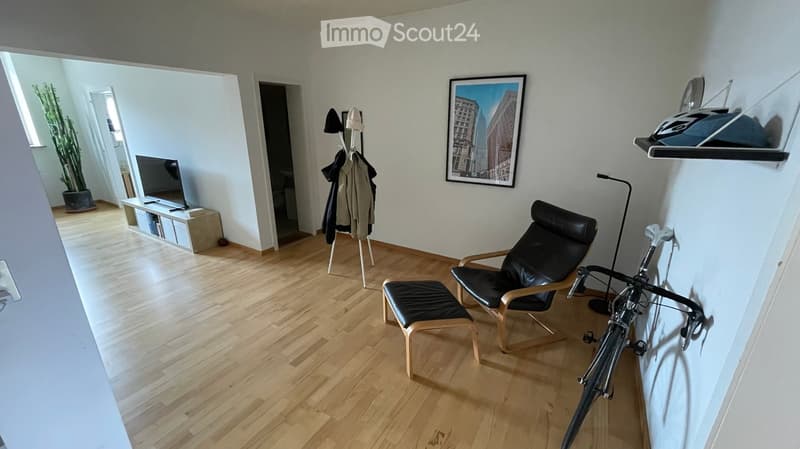 A modern living room with wooden flooring, a black leather chair, a matching stool, a television on a stand, a coat rack with jackets, a bicycle leaning against the wall, and a large picture frame on the wall.
