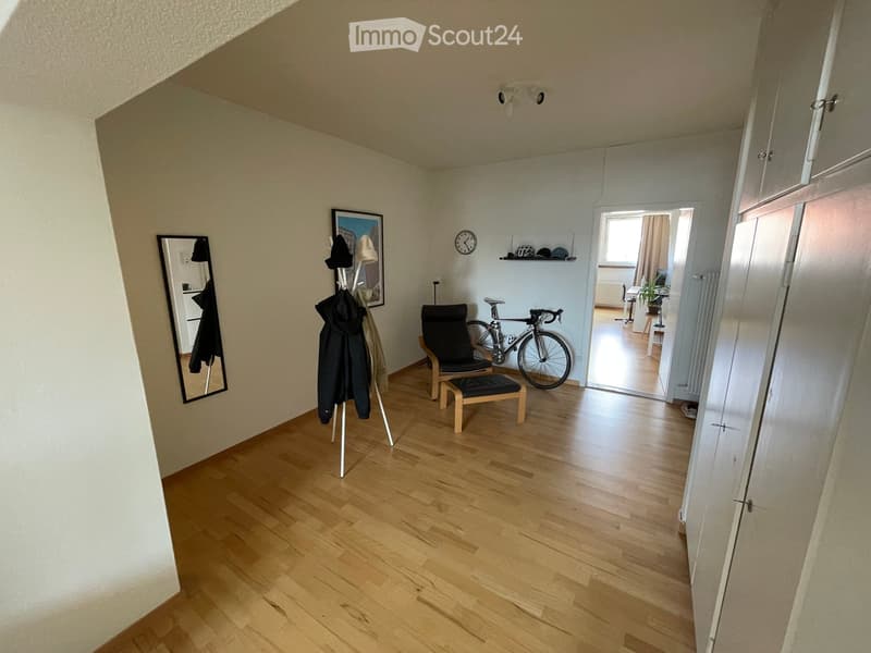 A small, well-lit room with wooden flooring, a chair, a stool, a bicycle, a mirror, a clock, a shelf, and a coat rack.