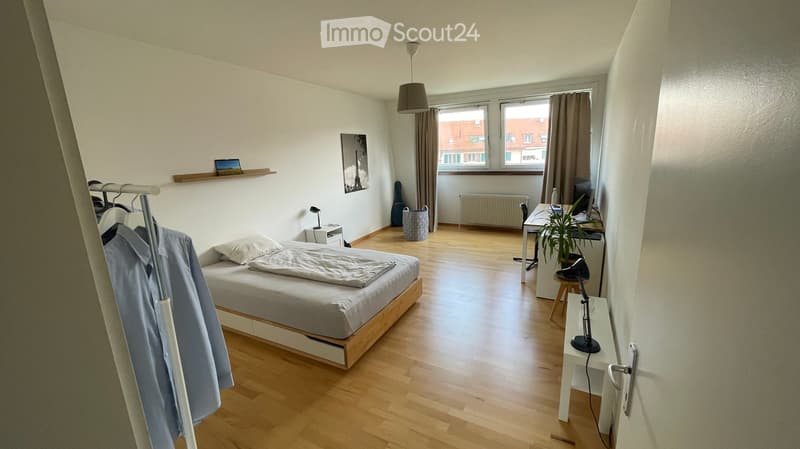 A modern, well-lit bedroom with wooden flooring, a single bed, a desk with a chair, a lamp, a plant, and a guitar leaning against the wall. The room features large windows with curtains and a shelf with a picture frame.