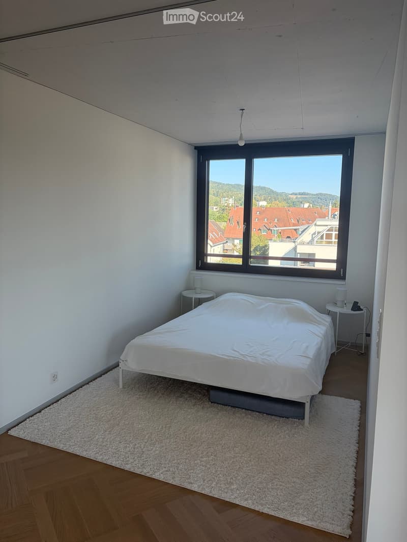 Minimalist bedroom with a white bed, nightstands, table lamps, and a large window overlooking a cityscape.