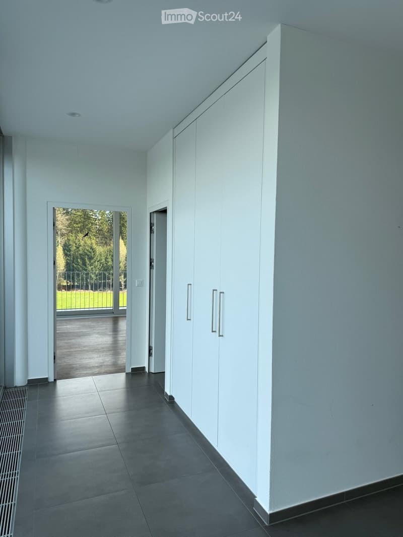 Modern hallway with white walls, dark tiled floor, built-in white storage units, and an open doorway leading to a balcony area with outdoor decking and greenery visible.