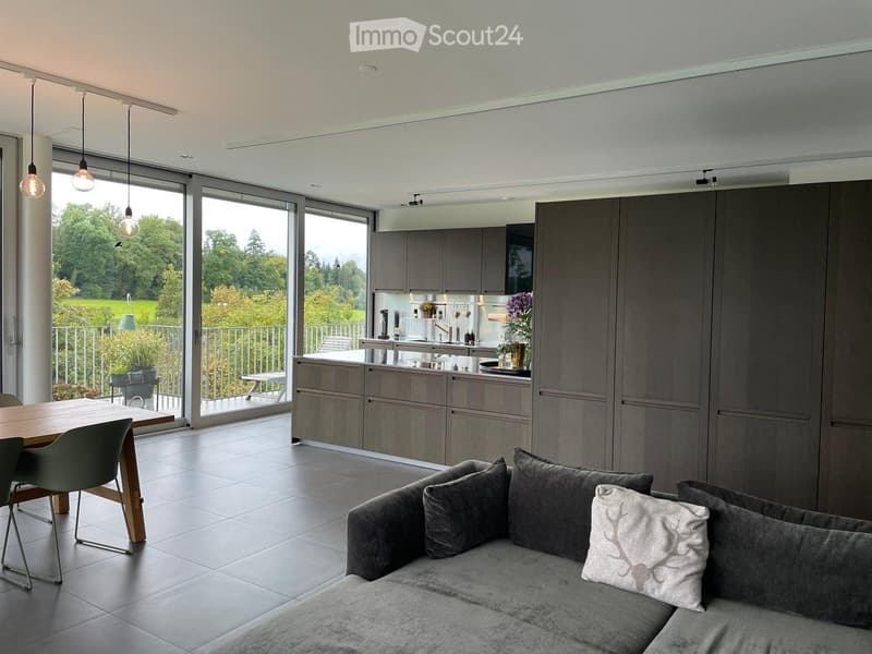 Modern open-plan living and dining area with grey sofa, dining table, and kitchen area featuring grey cabinets and a central island. Large windows provide a view of a green landscape.