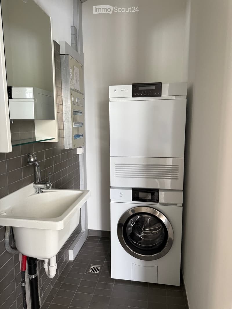 Modern bathroom with a white sink, chrome faucet, and a stacked washing machine and dryer unit. The walls are tiled in dark gray, and there is a small mirror and cabinet above the sink.