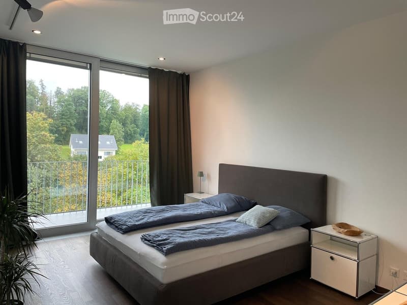 Modern bedroom with a large bed, nightstand, lamp, and a view of a green landscape through large windows.
