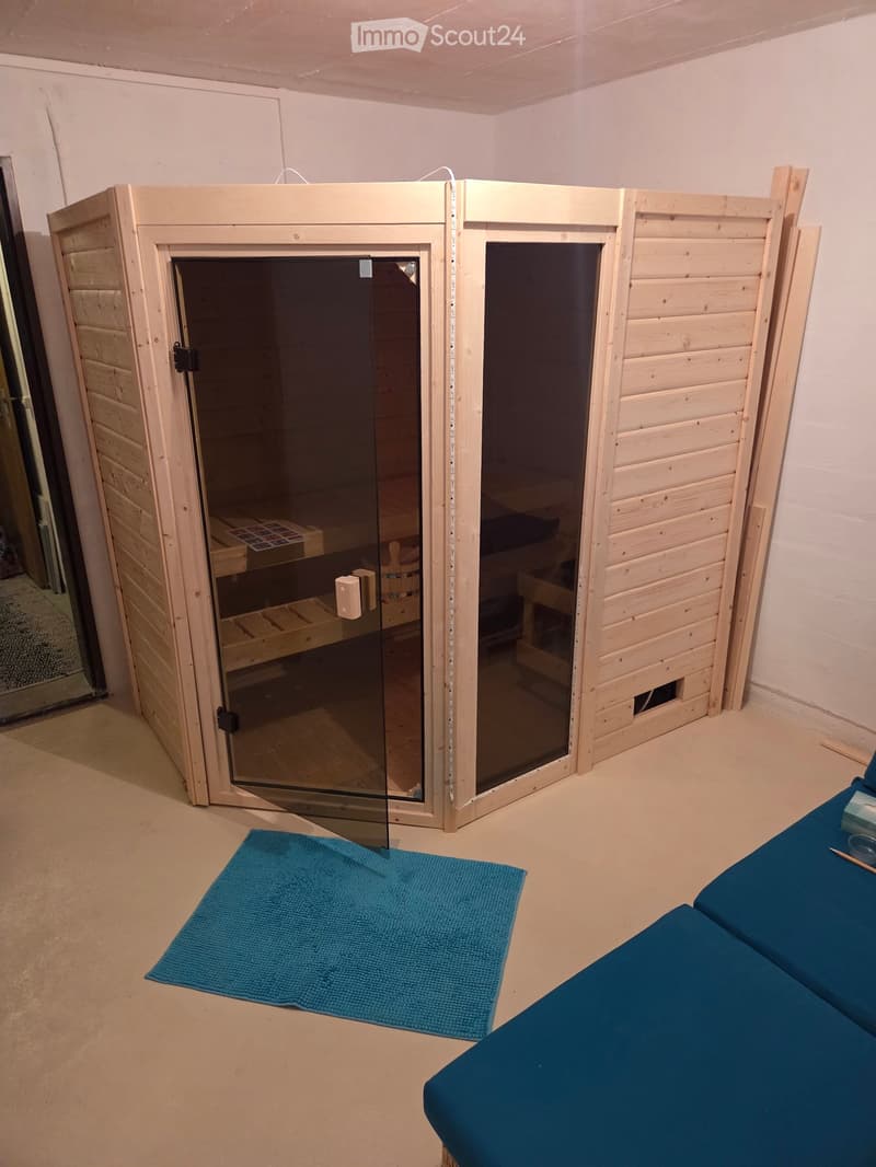 A small, well-lit room with a wooden sauna structure featuring glass doors, a blue towel on the floor, and a blue sofa in the corner.