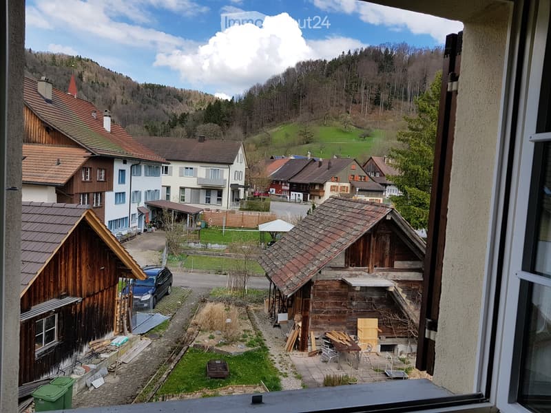 View from a window showing a rural village with traditional wooden houses, a small garden area with a table and chairs, and a parked car.