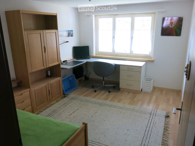 Well-lit room with a desk setup, a bed, and a large window letting in natural light. The room features a wooden cabinet, a computer, a chair, and a colorful artwork on the wall.