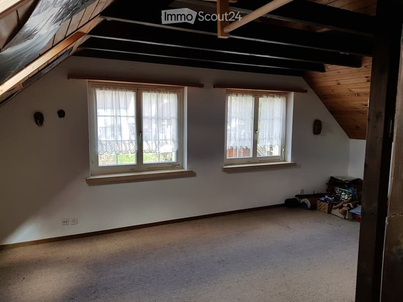 Empty attic room with wooden beams, skylight, and two windows with white curtains. Bare walls and carpeted floor.