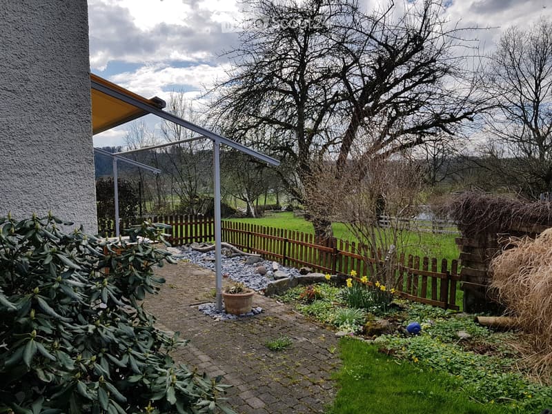Garden area with a paved pathway, wooden fence, potted plants, and a covered patio area attached to a building.