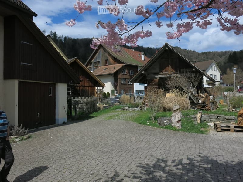A rural garden area with a paved brick pathway, wooden houses, a cherry blossom tree, and scattered garden items like a wooden pallet and stone sculptures.