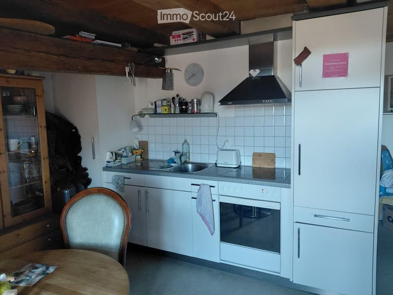 Modern kitchen with white cabinets, stainless steel appliances, and a wooden dining area. Features include a refrigerator, oven, sink, microwave, and a wooden dining table with a chair.