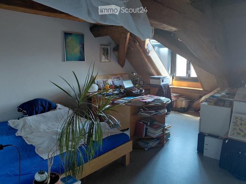 Cozy attic bedroom with slanted wooden ceilings, a bed with blue bedding, a desk cluttered with books and a laptop, and a potted plant near the bed.