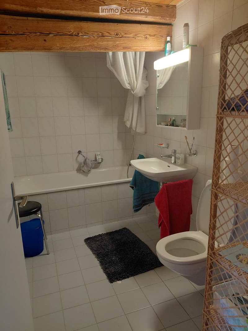 Bathroom with a bathtub, sink, toilet, mirror, and a small stool. Towels and a bath mat are visible, along with a toothbrush holder and a towel rack.
