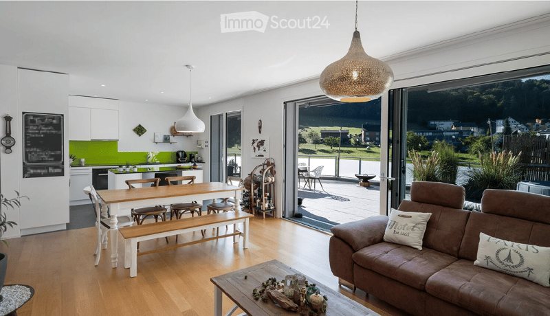 Open-plan living and dining area with modern kitchen, large windows offering a scenic view, and contemporary furniture including a sofa, coffee table, dining table, and kitchen appliances.