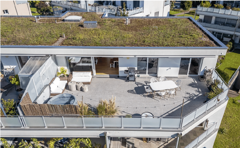 Modern rooftop terrace with a hot tub, seating area, dining table, and barbecue, surrounded by greenery and glass railings.