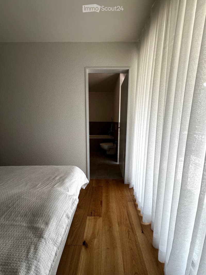 Bedroom with wooden flooring, a neatly made bed on the left, and an open doorway leading to a small bathroom with a toilet and sink.