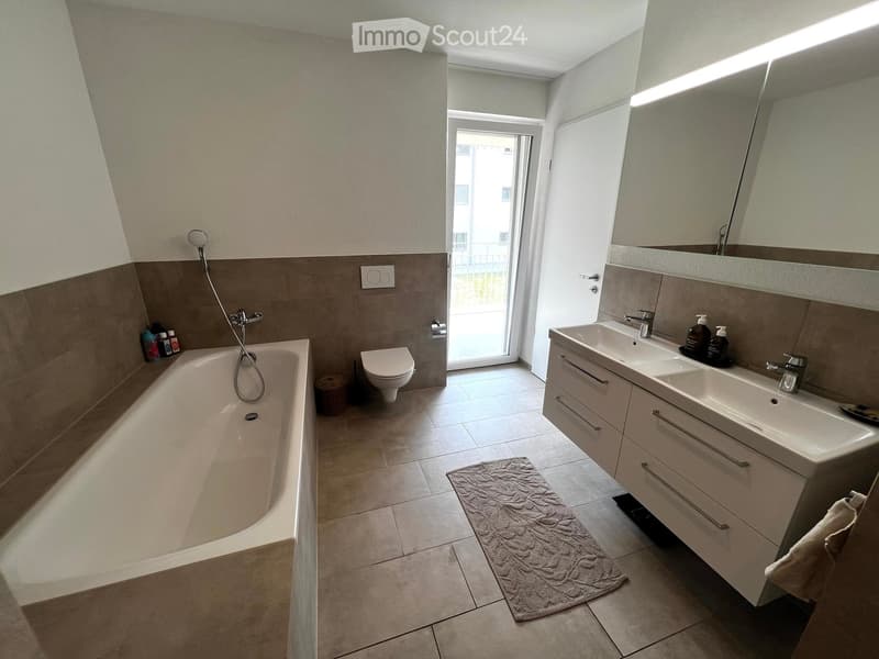Modern bathroom with a bathtub, toilet, double sink, large mirror, and a small window leading to a balcony.