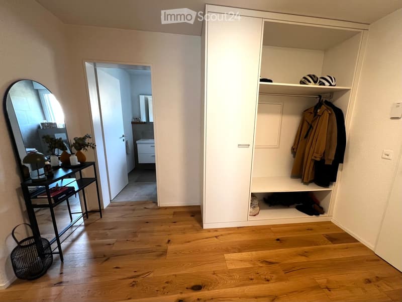 A well-lit hallway with wooden flooring, a large built-in wardrobe, a small table with a lamp and vases, and an open doorway leading to a bathroom.