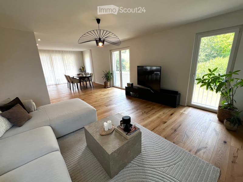 Modern living room with a white sectional sofa, coffee table, TV, dining area, and large windows with sheer curtains.