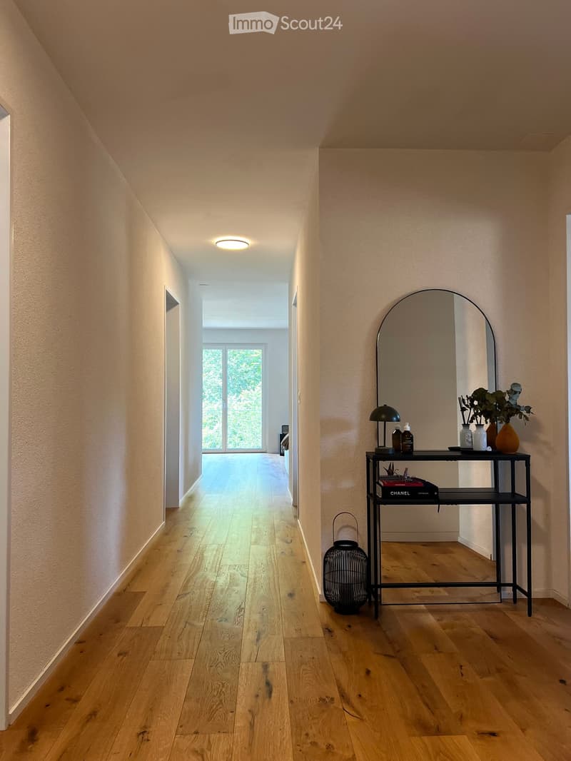 A modern hallway with light wooden flooring, beige walls, and a sleek black console table holding decorative items, a mirror, and a lamp.