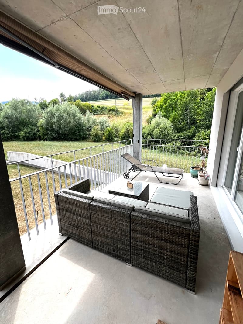 Balcony with a modern design, featuring a wicker sofa, a glass table, a lounge chair, and potted plants, overlooking a green landscape.
