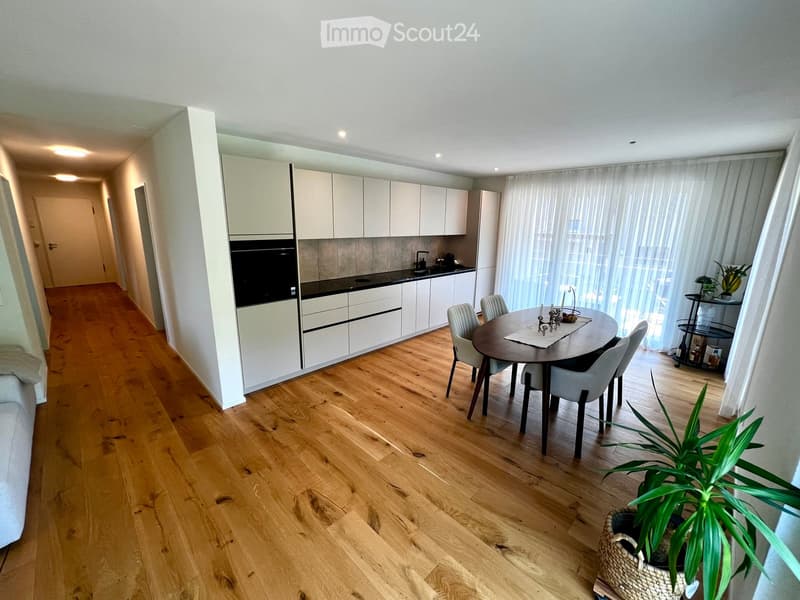 Open-plan kitchen and dining area with light wood flooring, white cabinets, a round dining table with chairs, and a potted plant near the window.