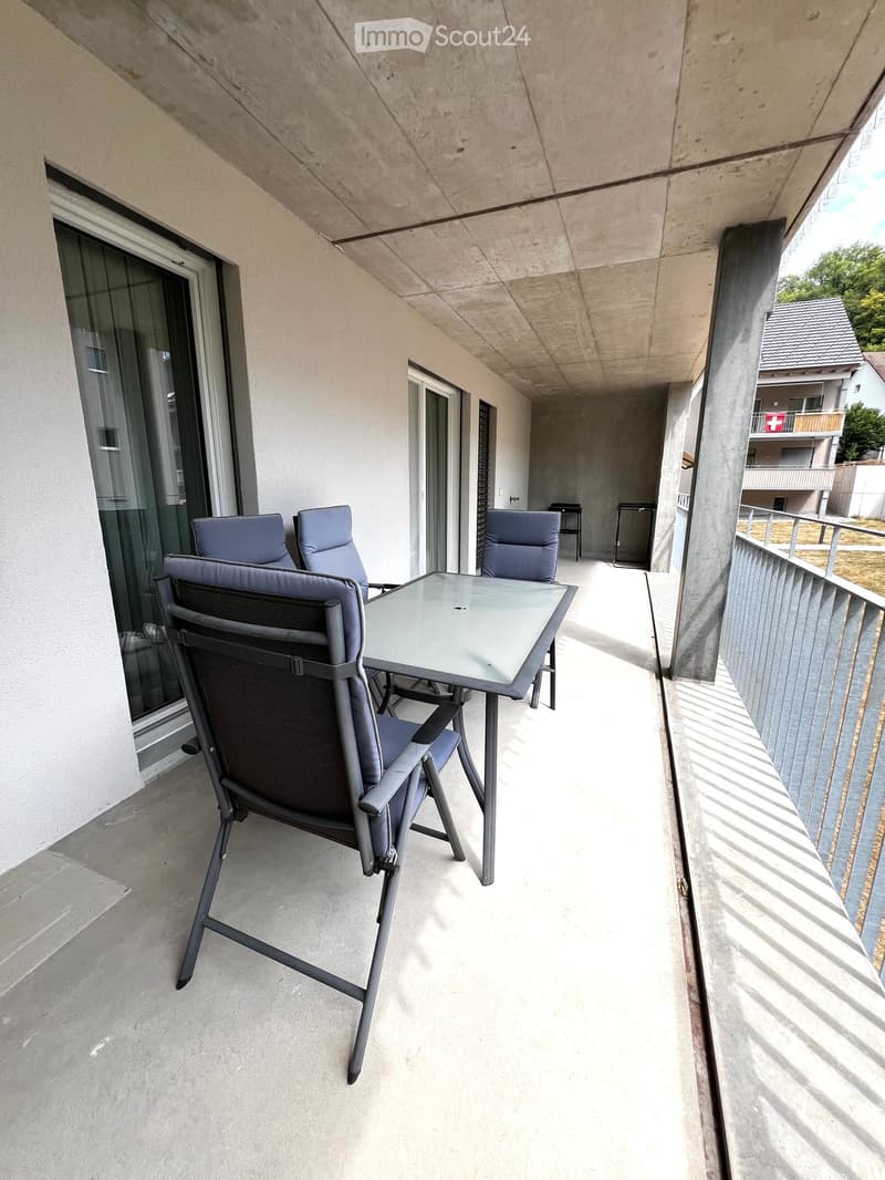 Balcony with a glass table and four chairs, concrete floor, and a view of neighboring buildings.