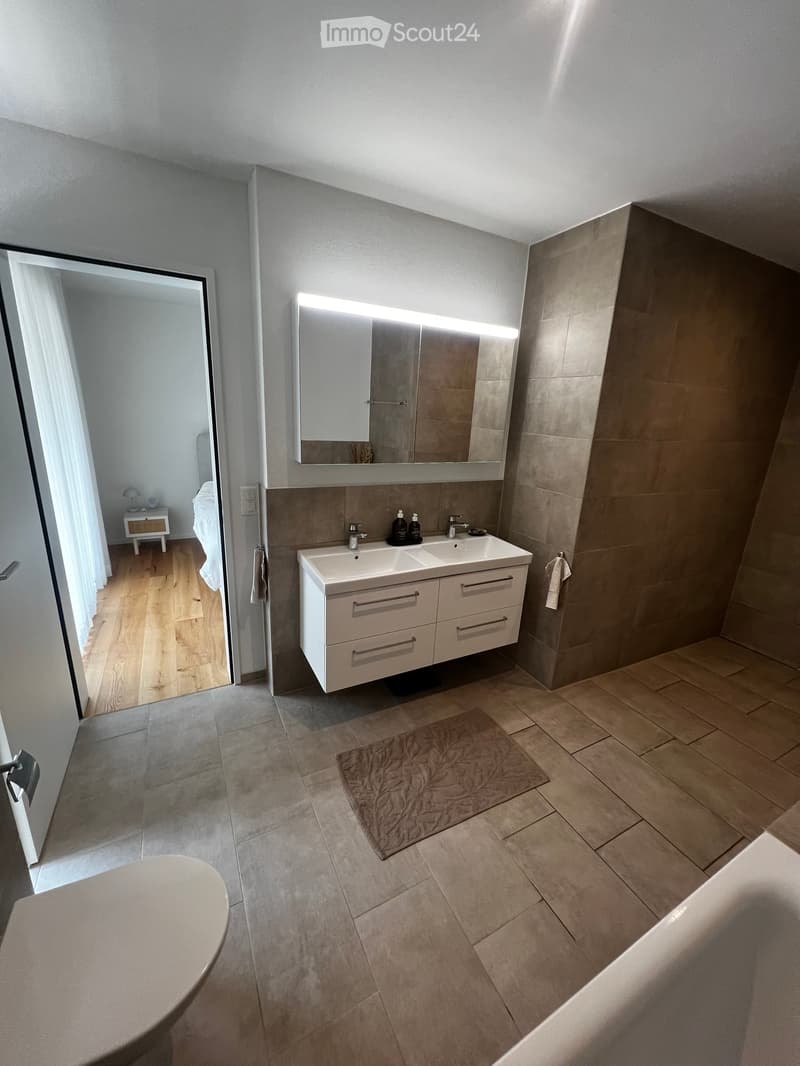 Modern bathroom with a double sink, large mirror, built-in cabinet, and a bathtub. The room features tiled floors and walls, a small rug, and an open doorway leading to a bedroom area with wooden flooring and a small nightstand.