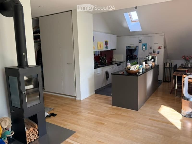 Modern kitchen with light wooden flooring, white cabinets, a black kitchen island, and a wood-burning stove. The space includes a sink, stove, fridge, and a dining area with a table and chairs.