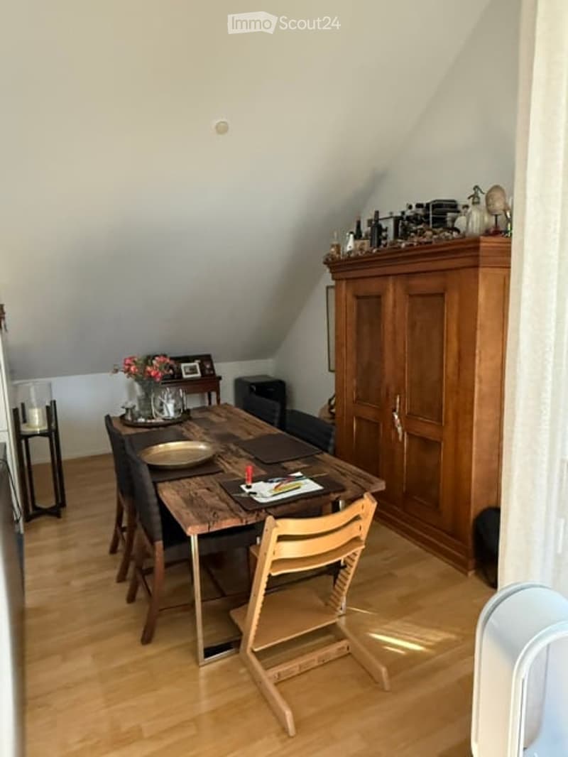 A cozy attic dining area with wooden floors, a large wooden dining table surrounded by chairs, a tall wooden cabinet, and decorative items on top.