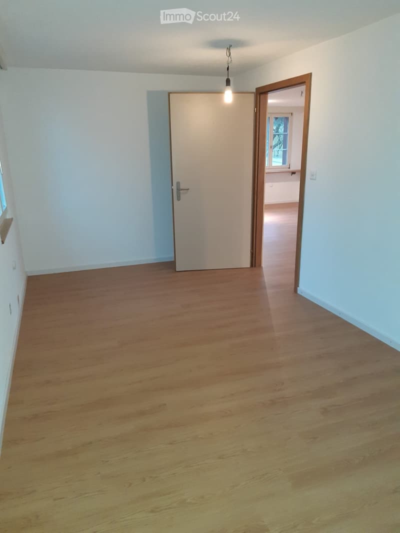 Empty hallway with wooden flooring, white walls, and two doors, one leading to another room with visible window and light fixture.