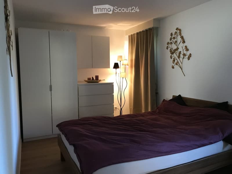 Modern bedroom with a wooden bed, dark red bedding, white wardrobe, nightstand with lamp, and decorative wall piece.