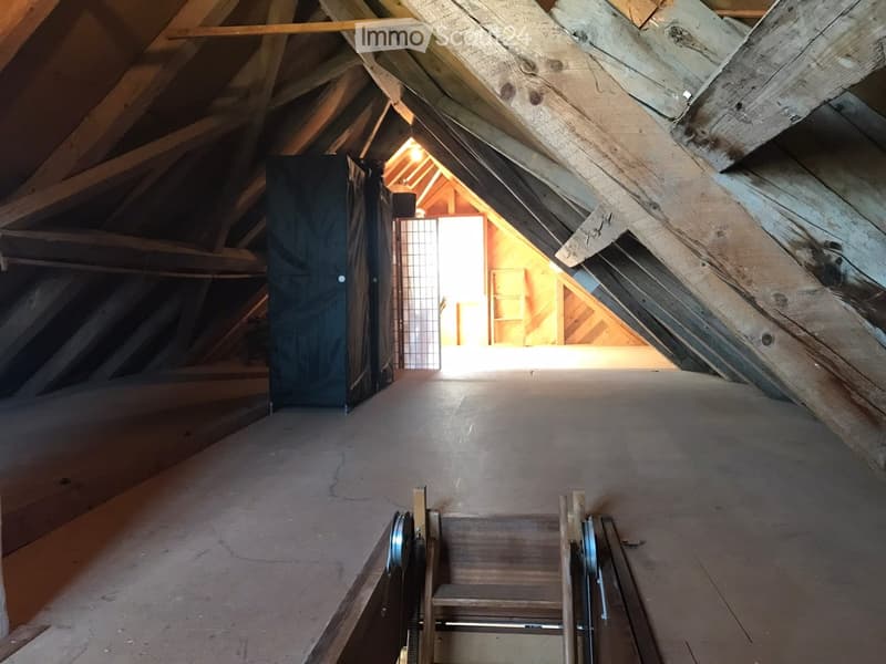 Empty attic space with wooden beams, exposed rafters, and a small opening to another room. The floor is unfinished, and there is a small staircase leading up.