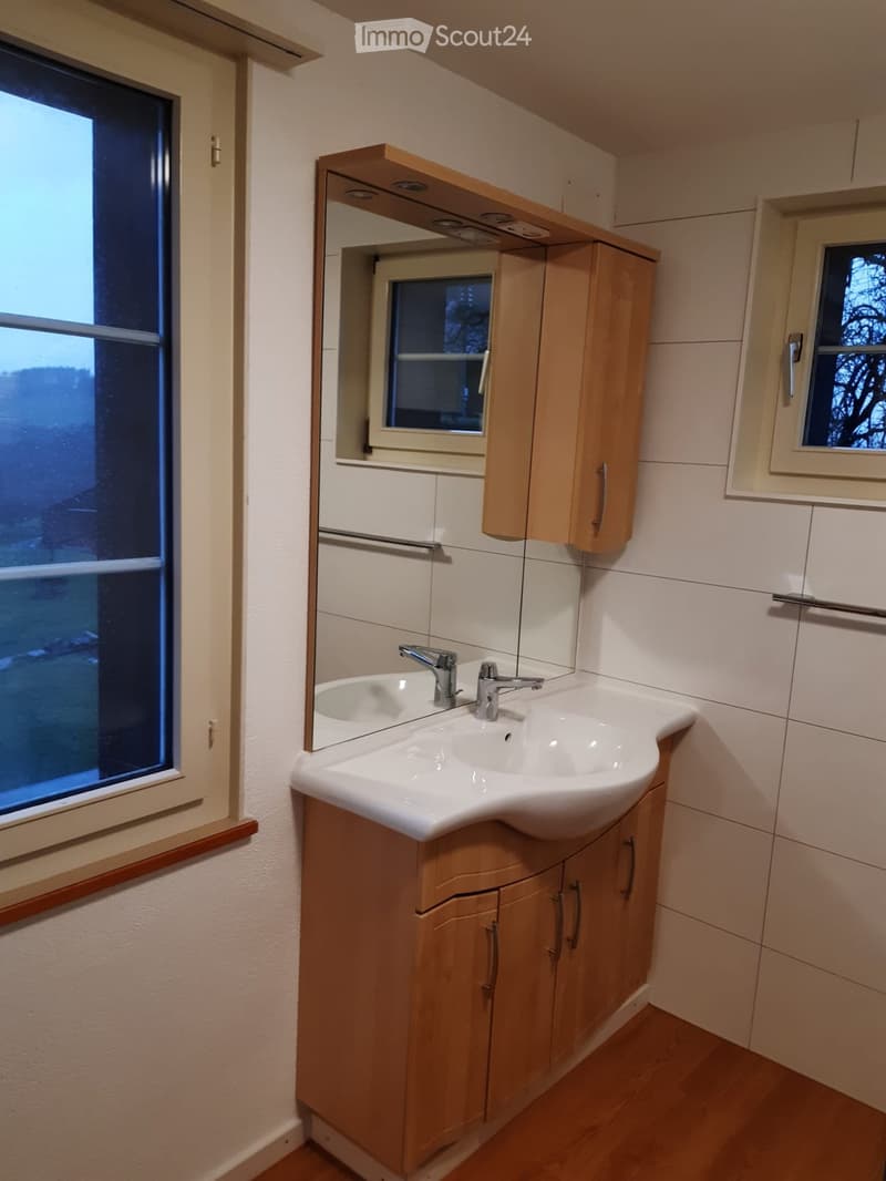 Modern bathroom with a white sink, wooden cabinet, and mirror above it. The room features light-colored tiles and a window providing natural light.