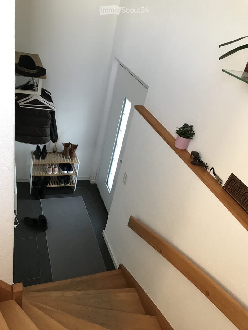 A modern hallway with wooden stairs, a small shelf for shoes, a coat rack, and a potted plant on the railing.