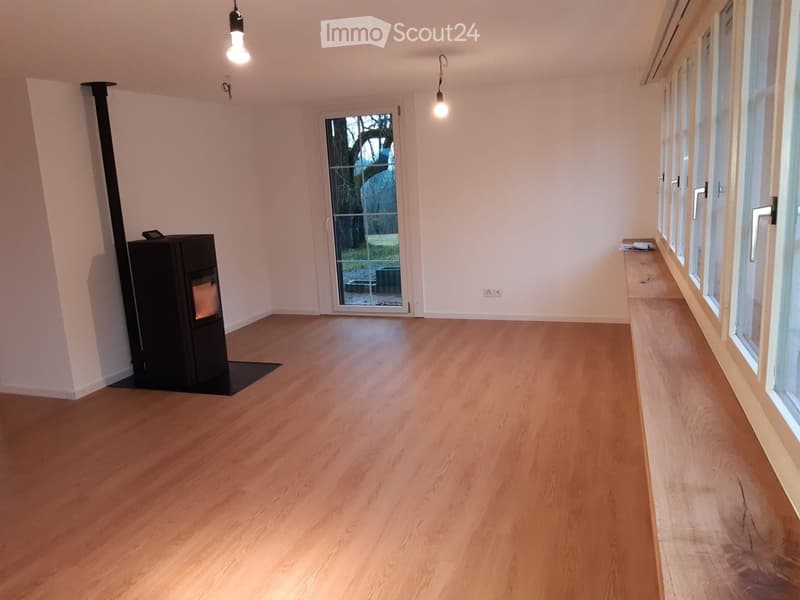 Empty room with light wooden flooring, white walls, large windows, and a black wood-burning stove.