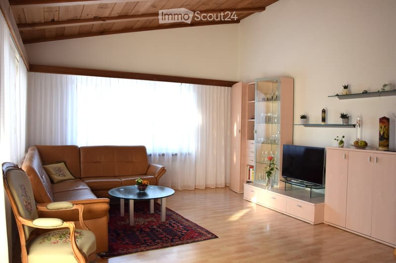 Living room with a brown leather sofa, a wooden armchair, a round coffee table, a television on a cabinet, shelves with decorative items, and a vase with flowers on the coffee table.