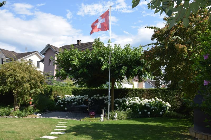 Well-maintained garden with lawn, flower beds, shrubs, trees, patio seating area, and Swiss flag on a pole.