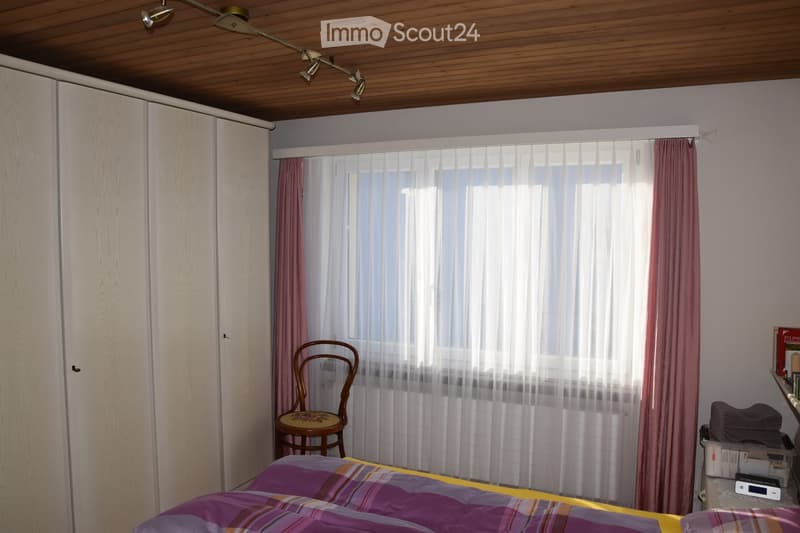 Bedroom with a bed covered in a purple and yellow striped duvet, a wooden chair with a floral seat, a large window with sheer white curtains and red side drapes, a white wardrobe, and a small table with folded clothes and a radio.