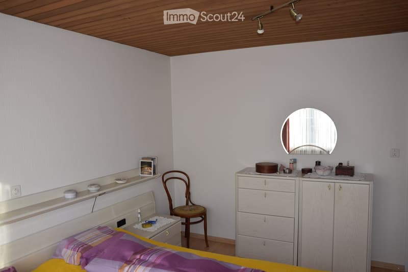 Cozy bedroom with a wooden bed, colorful bedding, a wooden chair, a vanity cabinet with a mirror, and various personal items on shelves and surfaces.