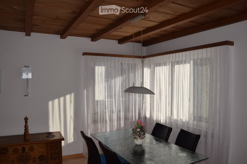 Bright dining area with a glass table, four black chairs, a flower vase with pink flowers, a wooden cabinet with a clock, and a hanging pendant light. Natural light streams in through sheer white curtains.