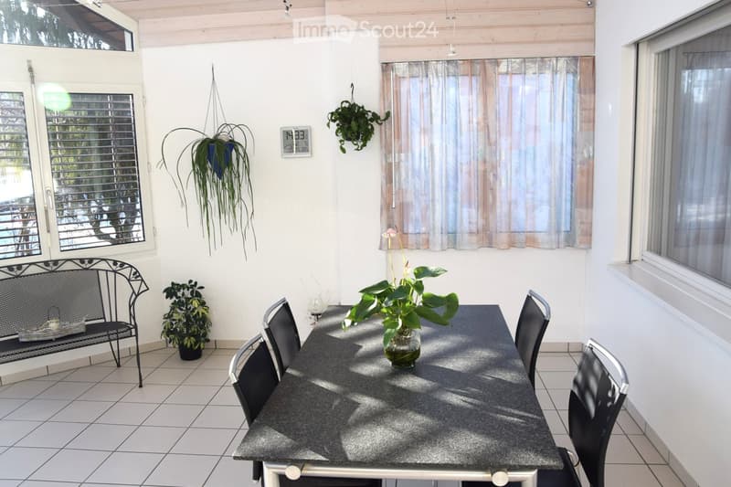 Balcony with tiled floor, white walls, large windows, a black metal bench, a dining table with chairs, and several potted plants.
